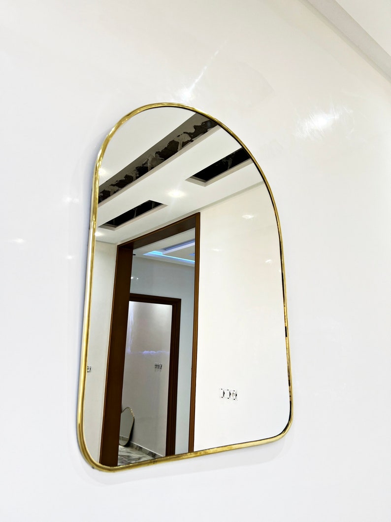 Puede incluir: Un espejo arqueado con marco dorado, parte superior redondeada y superficie lisa y reflectante. El espejo est&aacute; montado en una pared blanca y refleja un pasillo con una puerta de madera.