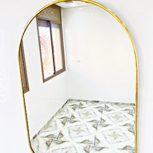 Puede incluir: Un espejo arqueado con marco dorado que refleja un suelo de baldosas blancas y una ventana con un panel de vidrio estampado.