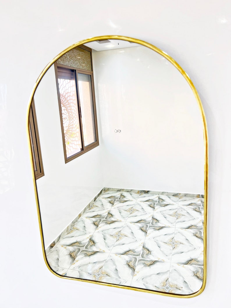 Puede incluir: Un espejo arqueado con marco dorado que refleja un suelo de baldosas blancas y una ventana con un panel de vidrio estampado.