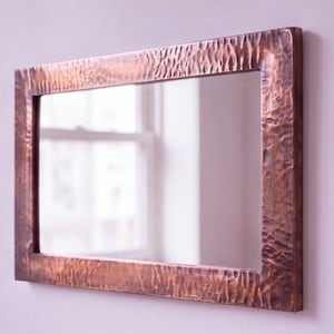 May include: A rectangular wall mirror featuring a wide, metallic copper frame with a distinct hammered, textured surface. The reflective glass surface is clear and smooth, showing a reflection of a window and light interior space.