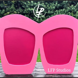 May include: A large pink, oversized sunglasses cutout, made of wood, with a white LFP Studios logo on the bottom.