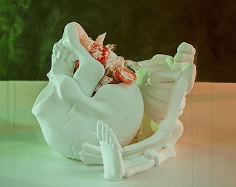 Pode incluir: Porta-doces de cerâmica branca em forma de esqueleto com uma tigela em forma de crânio cheia de doces embrulhados em vermelho e branco.
