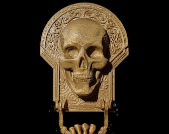 May include: A gold-colored skull door knocker with intricate details and a skeletal hand design. The knocker is mounted on a black background.