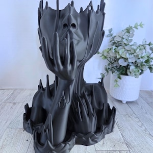 May include: A black decorative planter shaped like a human face, with the hands touching the face. The planter has a unique, artistic design with a textured surface and a modern aesthetic. The planter is suitable for small plants.
