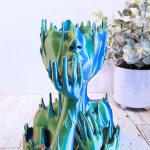 May include: A blue and green iridescent resin sculpture of a woman's head and torso with her hands clasped in front of her face. The sculpture is abstract and has a melted or dripping effect.