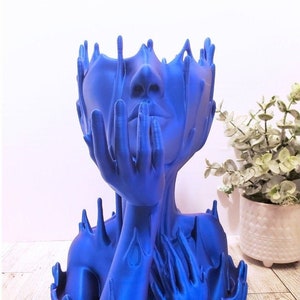 May include: A blue resin sculpture of a woman's head and shoulders with hands covering her face. The sculpture is abstract and has a flowing, liquid-like appearance.