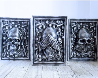 Pode incluir: Três peças de decoração de parede enquadradas em preto com um design de caveira. Cada caveira é cercada por um design de ossos e outros elementos esqueléticos. A caveira do meio está cobrindo os olhos com as mãos.