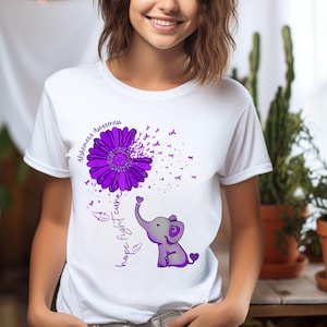 May include: White t-shirt with a purple flower and a purple ribbon design. The flower is a dandelion with purple ribbons blowing away. The text "Alzheimer's Awareness" is above the flower. Below the flower is a purple elephant with the text "Hope Fight Cure".