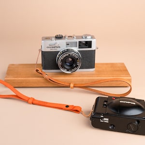 May include: Two vintage Olympus cameras, one silver and black 35 RC model and a black model, are displayed with an orange leather wrist strap. The 35 RC camera sits on a wooden block. The background is a solid beige color.