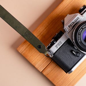 May include: A silver Nikon FE2 camera with a black leather grip and a green leather camera strap. The strap has the text "Click Support" printed on it. The camera rests on a wooden surface.