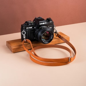 Puede incluir: Una cámara negra Minolta X-700 con lente, sobre un bloque de madera. Una correa de cuero marrón claro está en bucle por delante. La cámara tiene la palabra "Minolta" impresa.