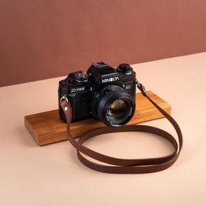 Puede incluir: Una cámara de película Minolta X-700 negra con una correa de cuero marrón descansa sobre un bloque de madera. La lente de la cámara está enfocada y la correa está en bucle. El fondo es un degradado de marrón y beige.