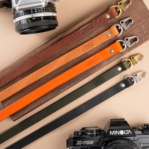 Puede incluir: Correas de cámara de cuero en varios colores, incluyendo marrón, naranja, verde y negro, con cierres metálicos. Las correas se muestran sobre una tabla de madera, con cámaras vintage en el fondo.
