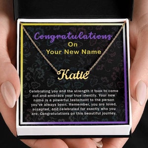 May include: A gold-colored necklace with the name "Katie" in cursive script, presented in a gift box. The box contains a card with the text "Congratulations On Your New Name". The card also includes a message about celebrating identity.