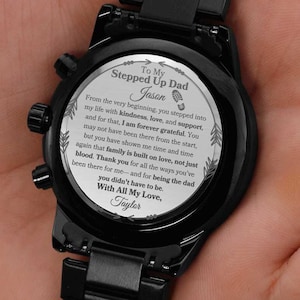 May include: Black wristwatch with a personalized message on the watch face. The message reads "To My Stepped Up Dad Jason" and includes a heartfelt message from "Taylor". The watch has a black metal band and casing.