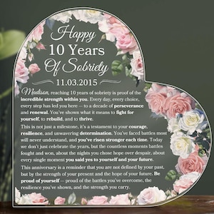 May include: A heart-shaped acrylic plaque with floral accents and text. The plaque reads "Happy 10 Years Of Sobriety" and includes a date. The design features pink and white roses.