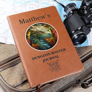 May include: A brown leather-bound journal with the text "Matthew's Dungeon Master Journal" on the cover. The journal features a circular image of a green dragon with red eyes and a floral background. A dice with crossed swords is also depicted on the cover.