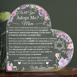Puede incluir: Una placa acrílica en forma de corazón con un diseño floral y el texto "Will You Adopt Me? Mom" y un mensaje sobre la familia. La placa también presenta un símbolo de triquetra, un símbolo de la Trinidad.