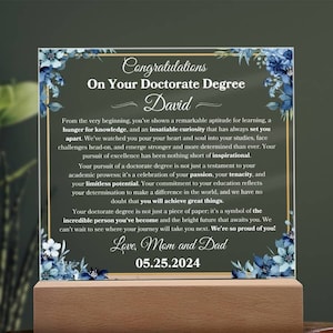 以下が含まれることがあります： 青い金色のフレームが付いたガラス製のプレートで、花柄のデザインと「博士号取得おめでとうデビッド」というテキストが書かれており、受賞者を称えるメッセージが書かれています。プレートには「05.25.2024」の日付と「愛を込めて、お母さんとパパより」という言葉も書かれています。