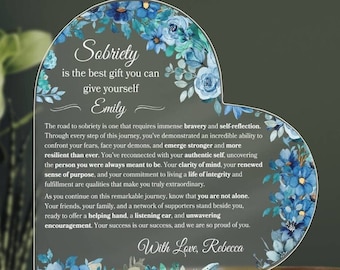 May include: A heart-shaped acrylic plaque with a blue floral design and a message about sobriety. The text reads "Sobriety is the best gift you can give yourself Emily. The road to sobriety is one that requires immense bravery and self-reflection. Through every step of this journey, you've demonstrated an incredible ability to confront your fears, face your demons, and emerge stronger and more resilient than ever. You've reconnected with your authentic self, uncovering the person you were always meant to be. Your clarity of mind, your renewed sense of purpose, and your commitment to living a life of integrity and fulfillment are qualities that make you truly extraordinary. As you continue on this remarkable journey, know that you are not alone. Your friends, your family, and a network of supporters stand beside you, ready to offer a helping hand, a listening ear, and unwavering encouragement. Your success is our success, and we are so proud of you. With Love, Rebecca"