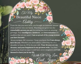 Puede incluir: Placa acrílica en forma de corazón con detalles florales y texto. La placa dice "Para mi hermosa sobrina Ashley" e incluye un mensaje de "Tía Joyce". Flores rosas y blancas adornan los bordes, creando un regalo decorativo.