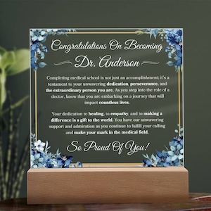 May include: A glass plaque with a gold frame and blue floral accents. The plaque has a message congratulating Dr. Anderson on becoming a doctor. The text reads "Congratulations on Becoming Dr. Anderson. Completing medical school is not just an accomplishment; it's a testament to your unwavering dedication, perseverance, and the extraordinary person you are. As you step into the role of a doctor, know that you are embarking on a journey that will impact countless lives. Your dedication to healing, to empathy, and to making a difference is a gift to the world. You have our unwavering support and admiration as you continue to fulfill your calling and make your mark in the medical field. So Proud Of You!"