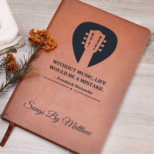 Könnte beinhalten: Ein braunes Lederjournal mit einer schwarzen Gitarren-Plektrumform und dem Zitat "Ohne Musik wäre das Leben ein Fehler. - Friedrich Nietzsche" und dem Text "Songs By Matthew" auf der Vorderseite.