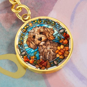 May include: A gold-toned keychain featuring a stained glass design of a brown and white dog with a blue and green background. The dog is surrounded by orange and yellow flowers.