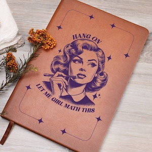 Könnte beinhalten: Ein braunes Lederjournal mit einer lila Illustration einer Frau mit einem Bleistift im Mund. Der Text "Hang On Let Me Girl Math This" ist auf dem Cover aufgedruckt.