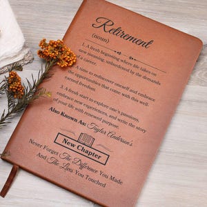May include: A brown leather journal with the word "Retirement" embossed on the cover. The journal includes definitions and inspirational text about retirement. The text "New Chapter" is also visible. A brown ribbon bookmark is attached.