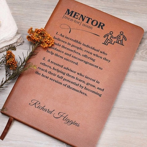 Puede incluir: Un diario de cuero marrón con la palabra "MENTOR" definida como "un individuo increíble que cree en la gente, incluso cuando dudan de sí mismos, ofreciendo orientación y aliento para ayudarlos a tener éxito". El diario está personalizado con el nombre "Richard Higgins".