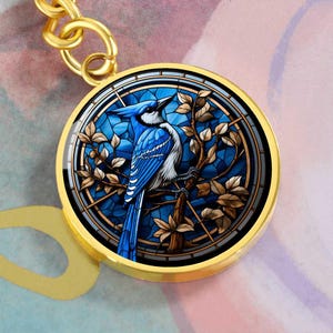 May include: A round, gold-colored pendant featuring a blue jay bird design. The bird is perched on a branch with brown leaves, set against a stained-glass style blue background. The pendant is attached to a gold chain.