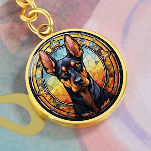 May include: A gold-toned keychain featuring a stained glass design of a black and tan Doberman Pinscher dog. The dog is depicted with a colourful background and intricate details.