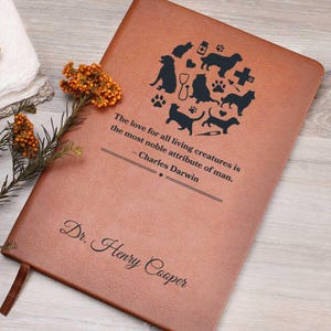 May include: A brown leather journal with a quote about the love for all living creatures by Charles Darwin. The journal is personalized with the name "Dr. Henry Cooper" in black lettering. The cover features a heart-shaped design with various animal silhouettes and veterinary symbols.