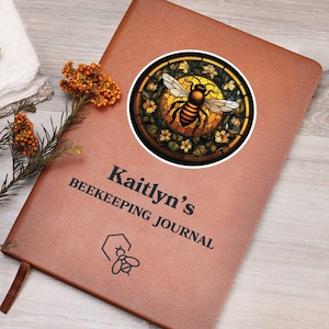 Puede incluir: Un diario de cuero marrón con el texto "Kaitlyn's Beekeeping Journal" y una pegatina de una abeja de vitral con un diseño de panal.
