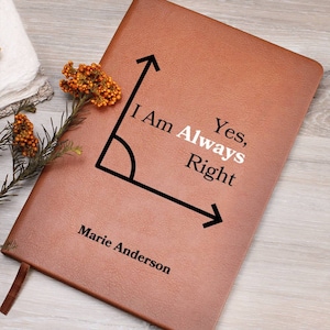 May include: A brown leather journal with the text "I Am Always Right" and "Marie Anderson" printed on the cover. A black angle graphic is also present. A brown ribbon bookmark is visible.