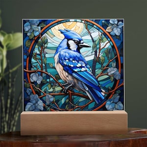 May include: A stained glass style artwork featuring a blue jay perched on a branch with blue and green leaves. The bird is surrounded by a circular frame with a sun-like design in the background.