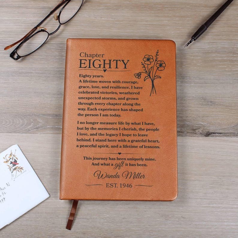 May include: A tan leather-like journal featuring the text Chapter EIGHTY, a floral illustration, and a personalized message for Wanda Miller, EST. 1946. The cover includes a ribbon bookmark, positioned near black eyeglasses and a black pen.
