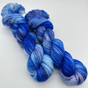 Könnte beinhalten: Zwei Stränge blaue Wolle mit weißen und violetten Streifen. Die Wolle ist diagonal angeordnet, wobei die Stränge verdreht und verknotet erscheinen. Die Farbpalette umfasst Blautöne, Weiß und Violett.