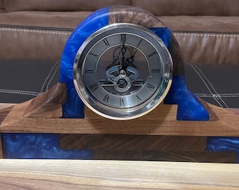 May include: A wooden clock with a blue resin base and a silver face. The clock has Roman numeral hour markers and a visible clockwork mechanism.