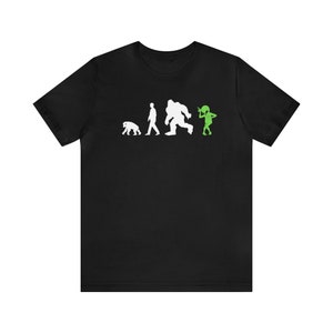 May include: Black t-shirt with a white graphic of the evolution of man, ending with a green alien. The alien is holding its head and looking to the side.