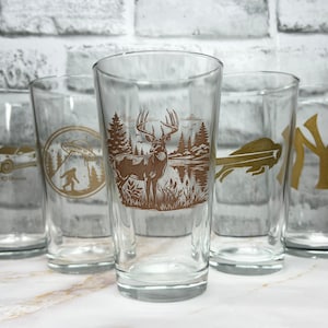 May include: Five clear glass pint glasses with gold designs. The designs include a UFO, a Bigfoot, a deer in the woods, a flying bird, and the New York Yankees logo.