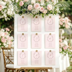 May include: A wedding seating plan featuring nine pink cards, each numbered and decorated with a floral wreath design. The cards are fixed to a white board, supported by a wooden easel. A floral arrangement of pink roses and white flowers sits above the board.