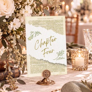 May include: A rectangular table number card featuring the text Chapter Four in gold script, layered over a vintage book page with torn edges. The card is held upright by a round, antique bronze-colored metal stand with a fleur-de-lis emblem.