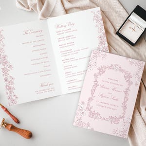 Op de afbeelding: Bruiloft briefpapier set met een roze bloemen design. Inclusief een trouwprogramma, uitnodiging en een ringdoosje. Een lakzegel en lepel zijn ook zichtbaar. De uitnodiging luidt "Welkom bij de bruiloft van Victoria Warner en Stephen Smith."