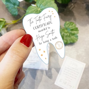May include: A white tooth-shaped certificate with blue edges, reading "The Tooth Fairy Certificate awarded to Hugo Smith for losing a tooth". It features gold stars and a gold seal with a tooth icon.