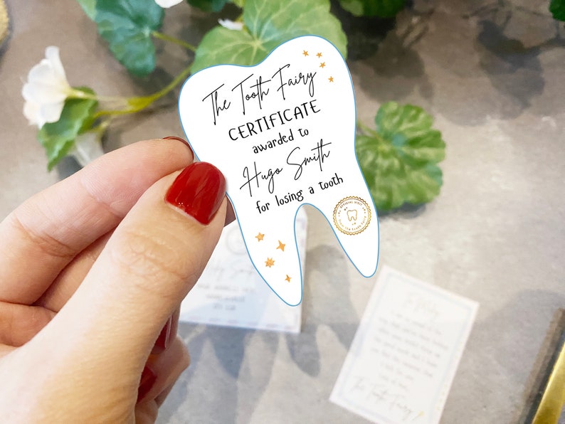 May include: A white tooth-shaped certificate with blue edges, reading "The Tooth Fairy Certificate awarded to Hugo Smith for losing a tooth". It features gold stars and a gold seal with a tooth icon.