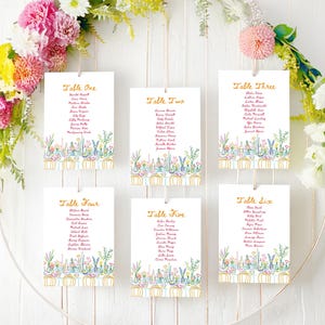 May include: Six white rectangular wedding seating chart cards with gold text headers Table One through Table Six, featuring colorful floral illustrations at the bottom, suspended by thin strings within a large circular floral wreath.