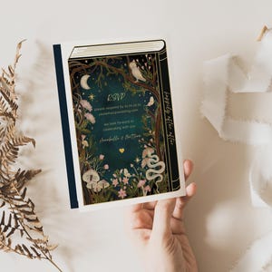 May include: A wedding invitation card with a dark blue background and gold accents. The card features a whimsical illustration of a forest scene with a book, flowers, and a snake. The text on the card reads "RSVP" and "happily ever after".