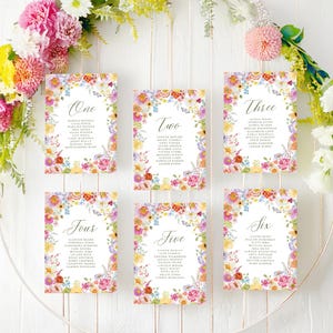 May include: Six rectangular wedding seating charts with floral borders in shades of pink, yellow, and purple. Each card features a large script number from one to six, followed by a list of names in a dark gray serif font, arranged on a circular frame.